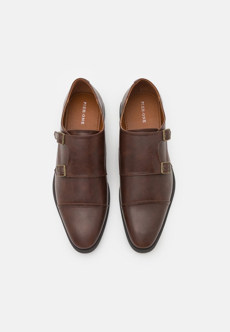 Scarpe da uomo in pelle marrone con due fibbie decorative, punta affusolata e suola nera a contrasto. Texture liscia con una finitura lucida.