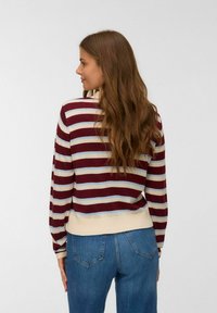 Vrouwen met lang golvend bruin haar, die een gemêleerd maroon, crèmekleurig en lichtblauw gestreept trui en blauwe jeans draagt, van opzij toont met de rug zichtbaar.