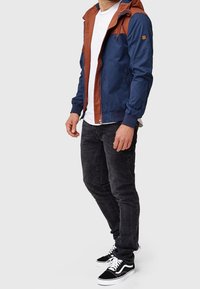 Homme portant une veste à capuche bleu marine et rouille, un t-shirt blanc, un jean noir et des baskets noir et blanc, debout devant un fond uni.