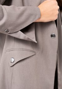 Light gray coat with a smooth texture, featuring large silver buttons and a flared collar. Notable side pocket with contrasting stitching detail.