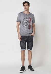 Camiseta gris con gráfico de motocicleta y texto, combinada con pantalones cortos de mezclilla negros con bolsillos laterales y dobladillos enrollados. Zapatillas negras completan el look.