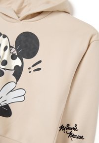 Beige Kapuzenpullover mit einem schwarzen und silbernen Minnie Mouse Grafik, weißen Akzenten und einer Unterschrift auf dem Ärmel. Weiches, lässiges Design.
