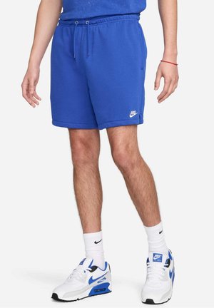 Hombre con pantalones cortos azules Nike, calcetines blancos Nike y zapatillas Nike Air Max blancas y azules, de pie frente a un fondo liso.