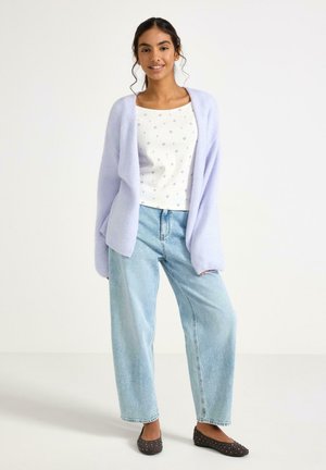 Nuori nainen seisoo hymyillen vaaleanvioletissa neulejakussa, valkoisessa, kuvioidussa neliskanttikauluspaidassa, leveälahkeisissa vaaleansinisisä farkuissa ja mustissa niiteillä varustetuissa ballerinoissa.