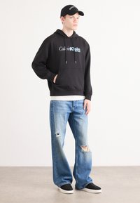 Black hooded sweatshirt with front pocket, featuring white and blue "Calvin Klein New York" text. Worn with light blue distressed jeans and black sneakers.