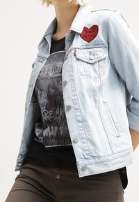 Hellblaue Jeansjacke mit einem Herz-Patch, ausgestattet mit bronzenen Knöpfen und zwei Brusttaschen, getragen über einem dunklen Grafik-T-Shirt.