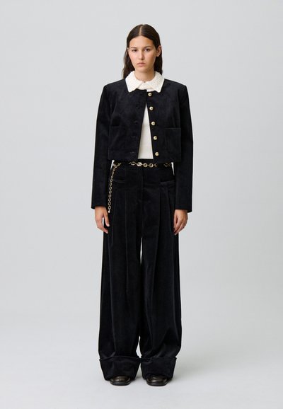 Giacca cropped nera in suede con bottoni dorati e tasche frontali, abbinata a pantaloni ampi neri in corduroy e un collare bianco testurizzato.