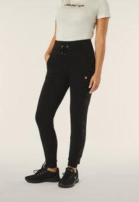 Pantalones de chándal negros con bolsillos laterales, cintura con cordón y detalles de encaje a lo largo del costado. Combinados con zapatillas deportivas negras.