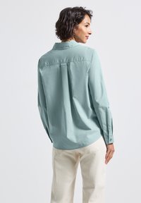 Una donna con capelli corti e scuri indossa una camicia a maniche lunghe di colore azzurro chiaro e pantaloni avorio, in piedi di spalle contro uno sfondo neutro.