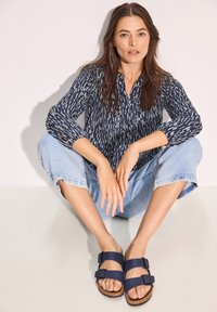 Blusa a fantasia navale con maniche lunghe, jeans cropped azzurri chiari e sandali navali con doppia fibbia. Tessuti morbidi e vestibilità rilassata.