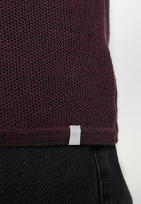 Suéter de punto burdeos con tejido texturizado, que presenta una etiqueta blanca de la marca en el dobladillo. El diseño incluye una banda ajustada en la parte inferior.