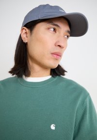 Green knitted sweater with a white logo on the chest, paired with a gray baseball cap. The cap features a slightly curved brim.