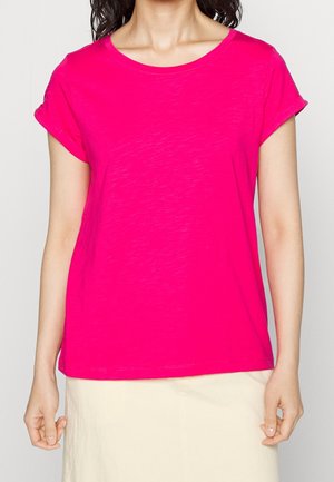 Vrouw draagt een felroze T-shirt met korte mouwen en opgerolde manchetten, en een crèmekleurige rok, staand tegen een effen achtergrond.