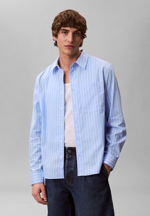 Junger Mann trägt ein hellblaues, vertikal gestreiftes Hemd über einem weißen Tanktop, kombiniert mit dunkelblauen Jeans, stehend vor einem schlichten Hintergrund.