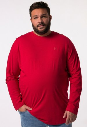 Rotes Langarmhemd aus weichem Material; mit Rundhalsausschnitt, strukturiertem Stoff und kleinem Logo auf der Brust. Getragen mit blauen Jeans.