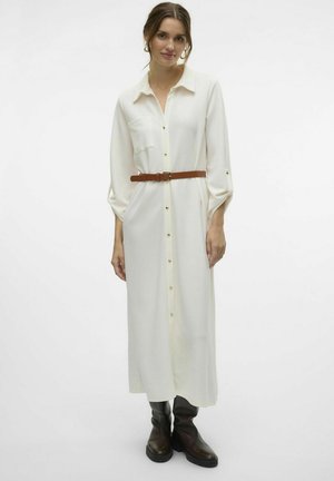 Lange, crèmekleurige shirtjurk met een kraag, gouden knopen, zijzakken en een bruine riem. Model draagt zwarte knie hoge laarzen.