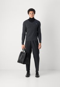 Sweter w kolorze węglowym z dekoltem w serek i długimi rękawami, połączony z czarnymi spodniami i czarnymi butami. Czarna skórzana torba w wzory trzymana w dłoni.