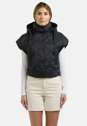 Frau trägt eine schwarze, gesteppte, ärmellose Kapuzenjacke über einem weißen Langarmshirt und hellbeigen Shorts, steht vor einem neutralen Hintergrund.