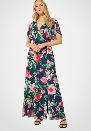 Kvinde iført en marineblå blomster maxi kjole med korte flagrende ærmer og sorte sandaler med hæl, der står med hænderne foldet.