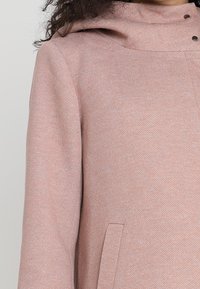 Ljusrosa kappa i en strukturerad tyg, med huva, två framfickor och tryckknappar. Designen har en minimalistisk och modern stil.