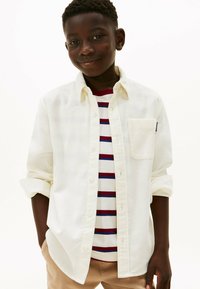 Camisa blanca de botonadura sobre una camiseta a rayas en rojo y azul marino. La camisa tiene un bolsillo en el pecho y las mangas arremangadas, combinada con pantalones beige.