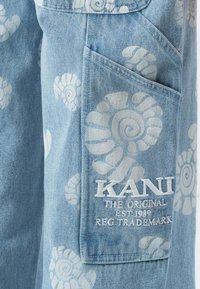 Világoskék farmer anyag fehér spirális kagylómotívumokkal és egy zsebével, amelyen az "KANI THE ORIGINAL EST. 1989 REG. TRADEMARK." felirat hímzett.