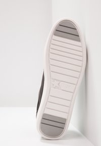 Semelle d'une basket grise avec une bande de roulement en caoutchouc blanc et gris, détail étoile près de la taille numéro 37, positionnée verticalement contre des murs blancs.