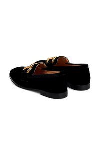 Par de mocasines de terciopelo negro con detalle de cadena dorada, tacón bajo y forro interior en tono tostado, mostrados desde una vista angular trasera.