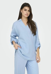 Chemise bleu clair à manches longues en V avec des poignets relevés, coupe décontractée, présentant des fentes latérales et des poches de chaque côté. Texture lisse.