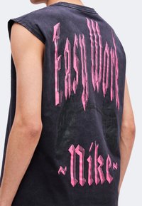 Personne portant un t-shirt noir sans manches avec un texte rose déformé et un motif noir délavé dans le dos sur un fond clair.