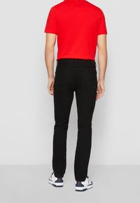 Jeans negros de corte ajustado con dos bolsillos traseros, combinados con una camiseta roja de manga corta y zapatillas blancas. Atuendo informal visto desde atrás.