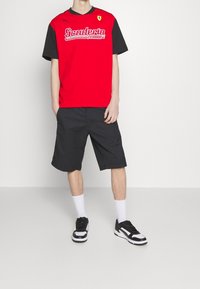 Camiseta de manga corta roja y negra con el logo de "Scuderia Ferrari", combinada con pantalones cortos negros. Se lleva con zapatillas negras y blancas y calcetines blancos.
