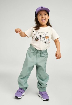 Light green elastic-waist pants, cream T-shirt with cartoon characters, and purple cap. Purple sneakers with velcro straps complete the outfit.