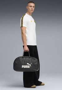 Mustast nahast duffelkott valge "PUMA" logoga, ümar kuju, topelt sangad ja sile tekstuur. Modell kannab valget särki mustriliste varrukatega.