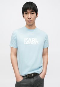 Világoskék pamut póló fehér "KARL LAGERFELD" logóval. Rövid ujjú, kerek nyakú. Sötét farmerrel és fekete övvel párosítva.