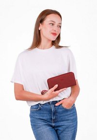 Red fabric wallet with a zip closure and a wrist strap, featuring a subtle texture and embossed logo. Held in a hand and paired with casual attire.