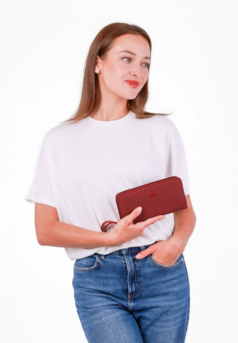 Red fabric wallet with a zip closure and a wrist strap, featuring a subtle texture and embossed logo. Held in a hand and paired with casual attire.