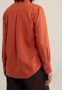 Rostfarbendes Hemd mit langen Ärmeln, strukturiertem Stoff, Rückenfalte und abgerundetem Saum, kombiniert mit dunklen Hosen.