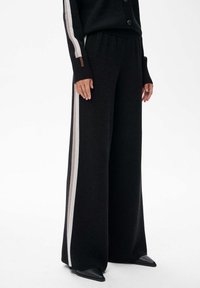 Czarne spodnie z szerokimi nogawkami i elastycznym pasem, z kontrastującymi białymi paskami po bokach i żebrowaną teksturą. Podkreślone małym logo.