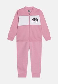 Survêtement rose en tissu doux, avec un panneau poitrine blanc et le logo "PUMA". Comprend une veste zippée et un pantalon assorti.