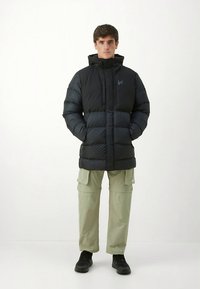 Fekete steppelt dzseki magas gallérral, elöl cipzárral és vízszintes varrásokkal; világoszöld cargo nadrággal és fekete tornacipővel viselve.