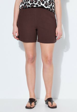 Vrouw draagt donkerbruine shorts, zwarte slip-on sandalen en een top met luipaardprint, staand tegen een witte muur.