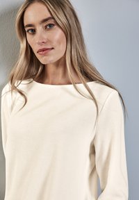 Top lungo a maniche con scollo a barca, di colore crema e texture, caratterizzato da una vestibilità leggermente rilassata e dettagli di cucitura visibili.