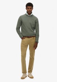 Suéter de punto verde con cuello, pantalones caqui y zapatillas crema. Presenta un emblema en el suéter y un cinturón marrón.