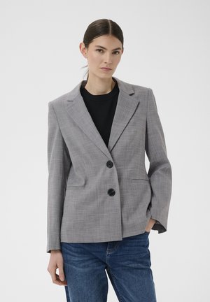 Frau mit zurückgebundenem dunklem Haar, die einen grauen karierten Blazer über einem schwarzen Oberteil und blauen Jeans vor einem schlichten Hintergrund trägt.