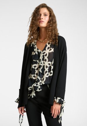 Cardigan nero sovrapposto a una blusa con volant che presenta un motivo geometrico bianco su sfondo nero. Maniche lunghe e decorate.