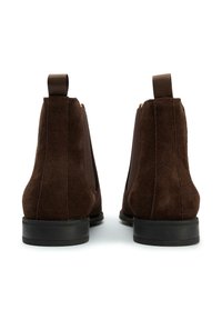 Brown suede Chelsea boots with elastic side panels and fabric pull tabs. Flat rubber sole with textured detailing. Simple and classic design.