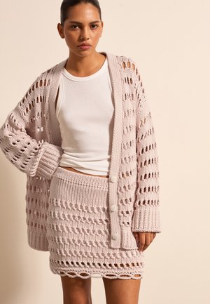 Helles rosa, gestricktes Cardigan mit offenem Strickdesign, langen Ärmeln und Knöpfen, kombiniert mit einem passenden, strukturierten Strickrock.