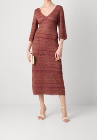 Robe tricotée bordeaux à longueur genou, avec un décolleté en V, des manches trois-quarts et une texture à motifs élaborés, associée à des sandales à talons beiges.