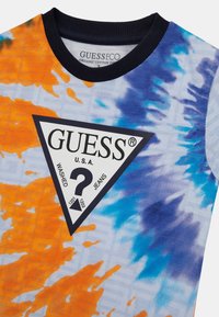 Koszulka z organicznej bawełny z wzorem tie-dye w kolorach pomarańczowym, niebieskim i fioletowym. Czarny, prążkowany dekolt z trójkątną metką logo.